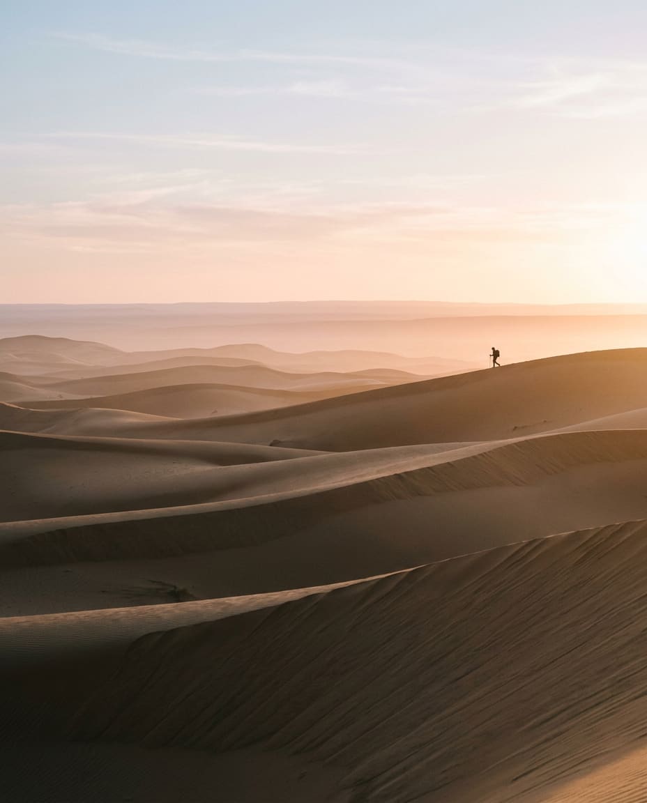 Silhouette d’un randonneur sur une crête, au milieu de dunes baignées de lumière rose à l’aube.